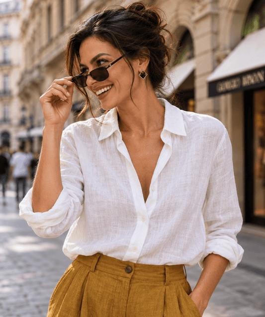 A woman wearing Nuno Felt earrings a white linen button-up shirt and mustard yellow linen trousers, standing on a cobblestone street in front of a European-style building.