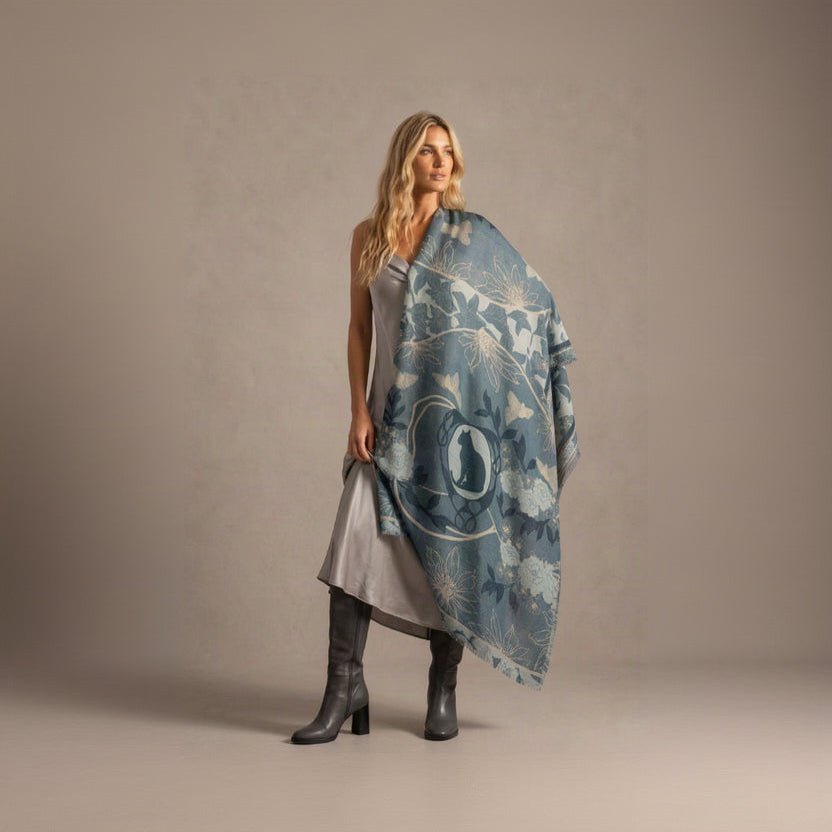 A blonde woman in a grey dress and grey knee-high boots, holding the Leopard Azure cashmere scarf open and draped over one shoulder like a cape, showing the leopard cameo, blue florals and fringed ends against a soft grey backdrop.
