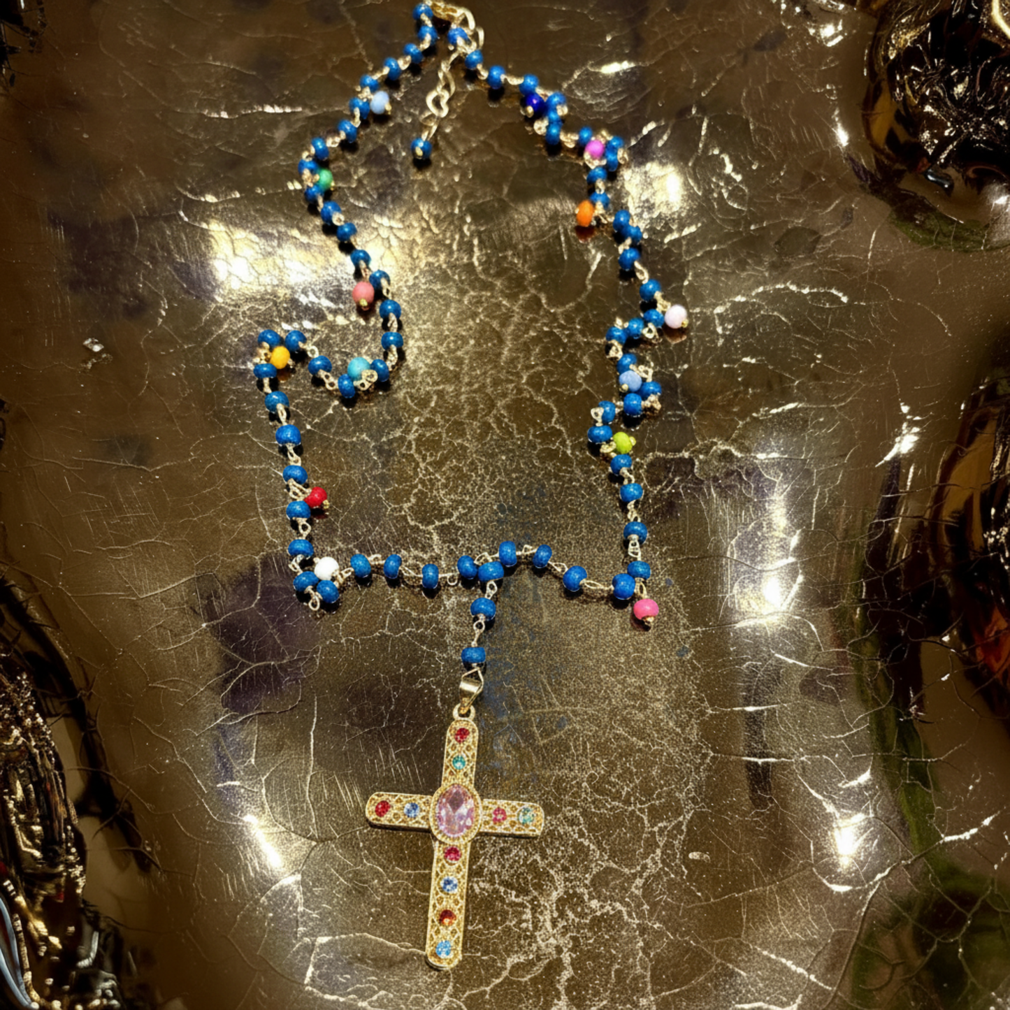 A beaded necklace with a gold-tone cross pendant, featuring predominantly blue beads with small multicolored accents, displayed on a reflective metallic surface.