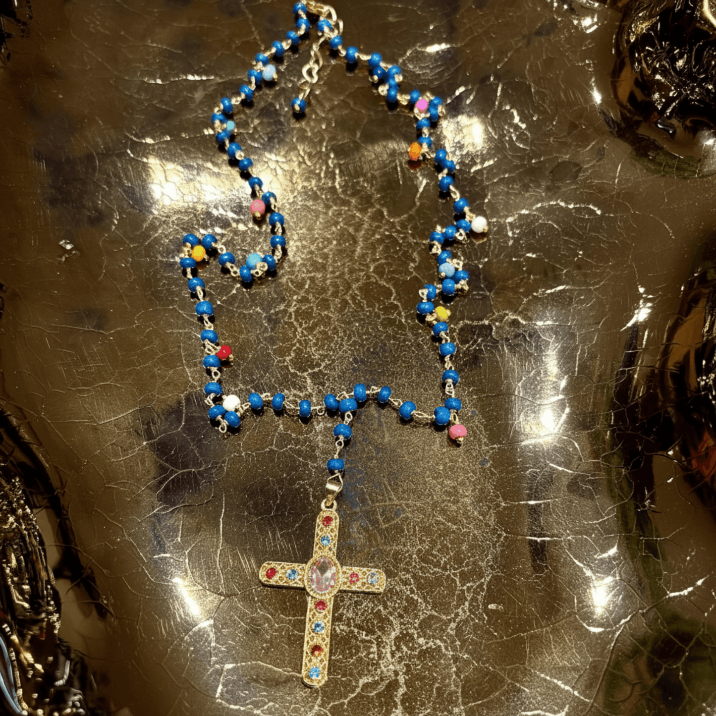 A beaded necklace with a gold-tone cross pendant, featuring predominantly blue beads with small multicolored accents, displayed on a reflective metallic surface.
