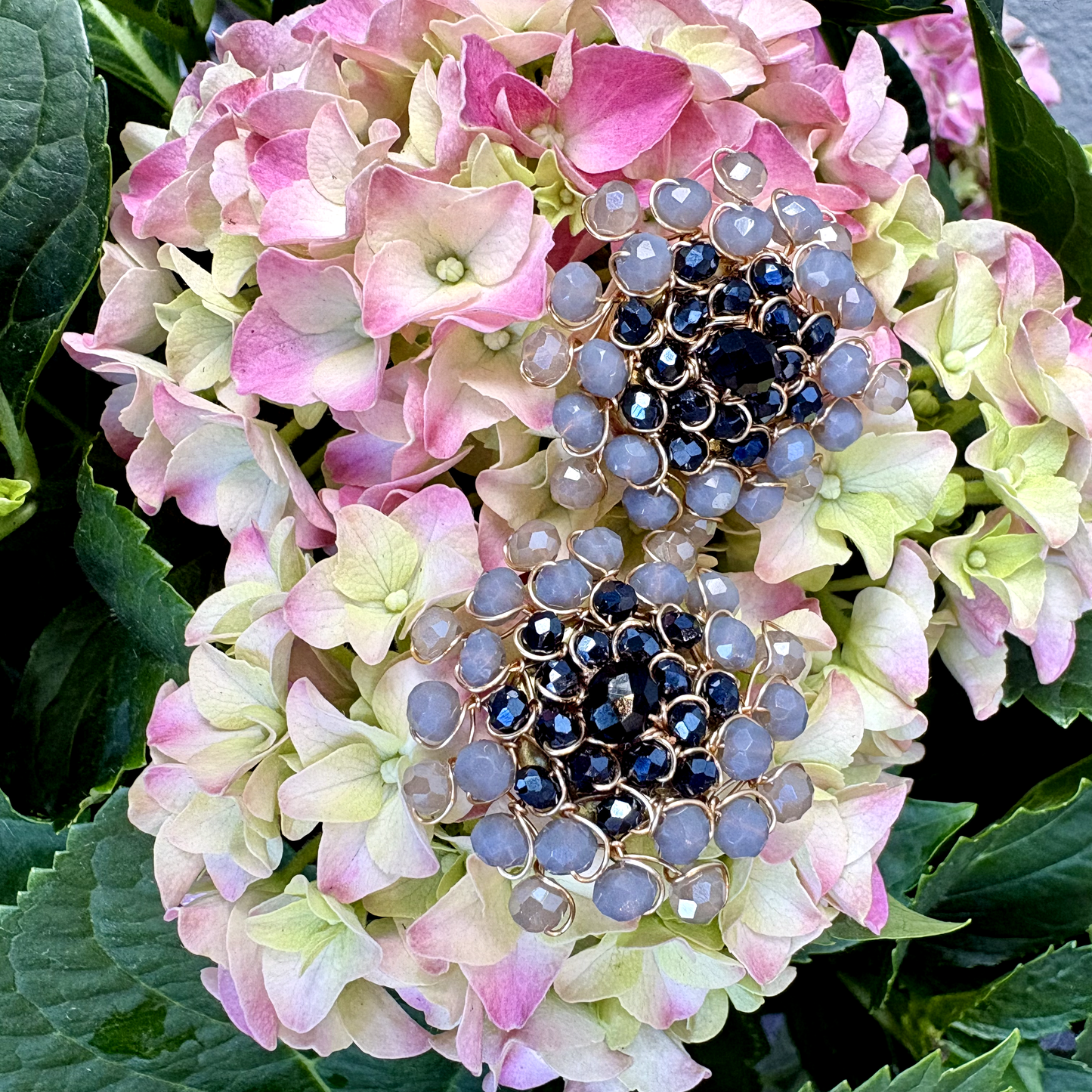 A pair of button covers featuring rows of faceted light blue-gray beads and dark blue center stones, displayed on a pink and green hydrangea flower.