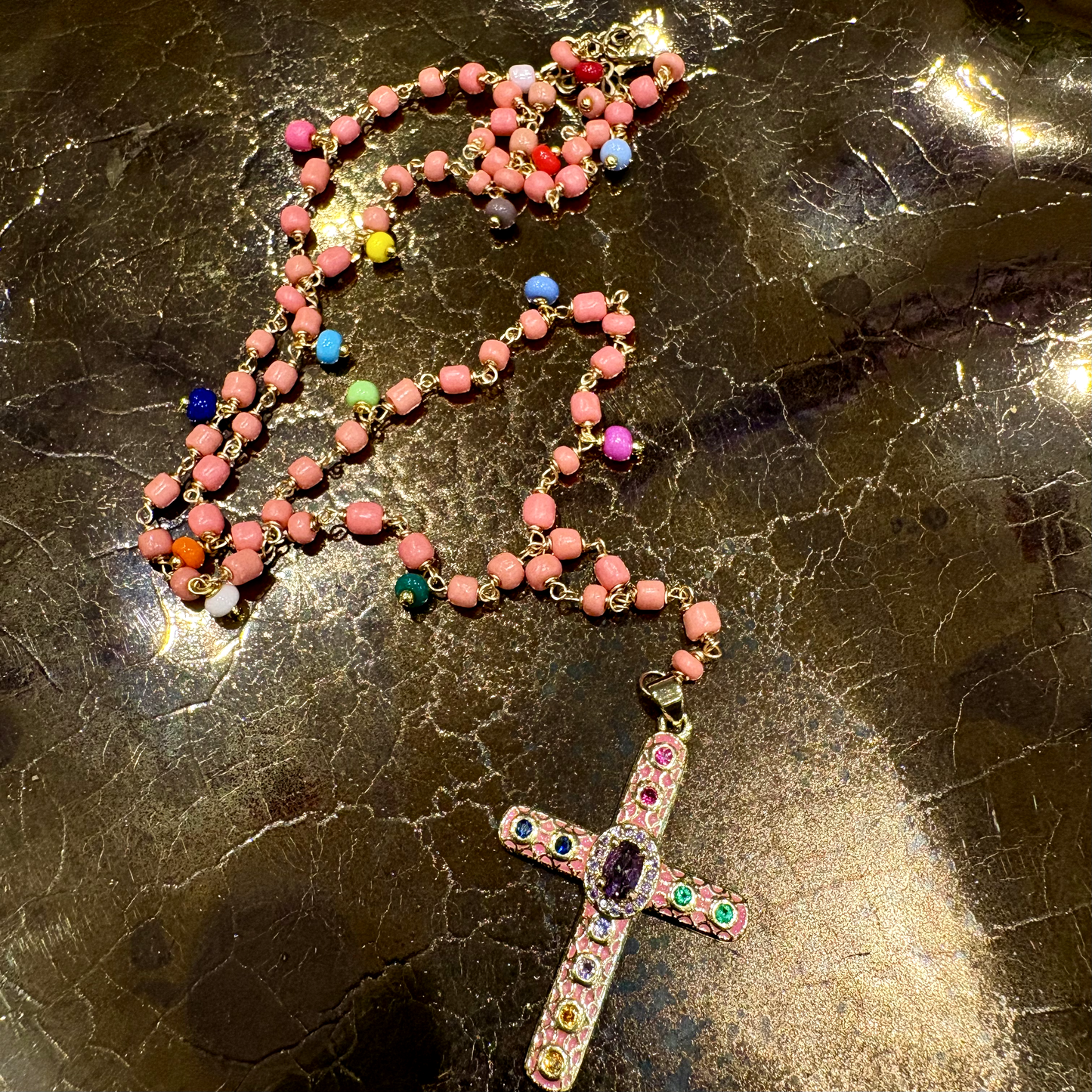 A rosary necklace with pinkish-red round beads and a detailed cross pendant, decorated with colorful rhinestones and small multicolored accent beads, displayed on a dark, glossy marble surface.