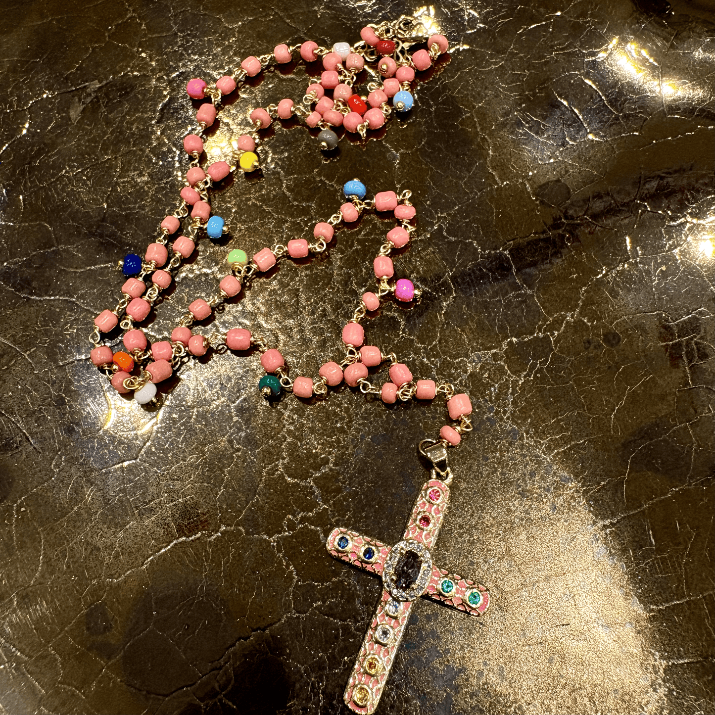 A rosary necklace with pinkish-red round beads and a detailed cross pendant, decorated with colorful rhinestones and small multicolored accent beads, displayed on a dark, glossy marble surface.