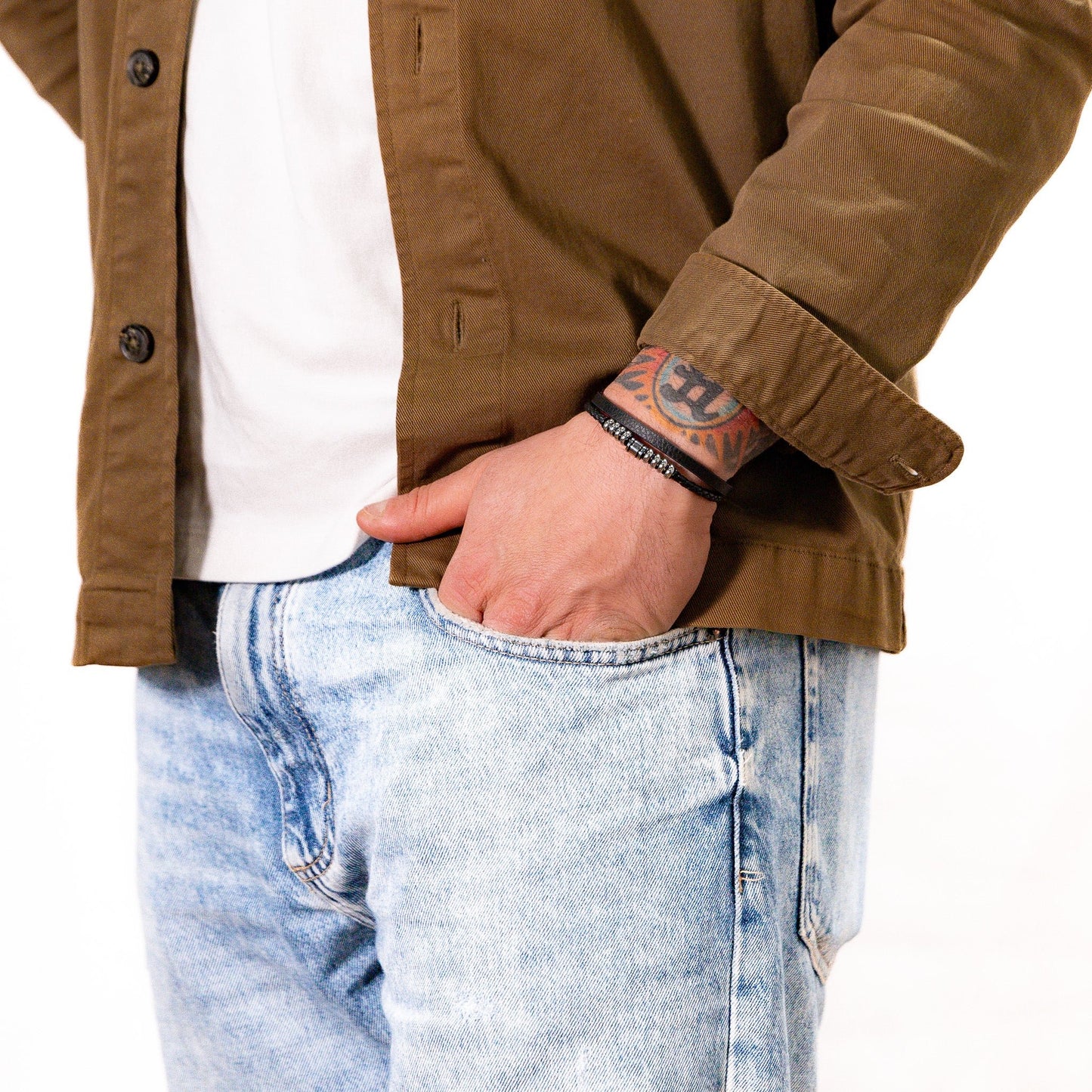 A man wearing a black double-strand leather bracelet with smooth and braided strands, polished silver stainless steel centre rings and magnetic snap closure with jeans and a brown over shirt.