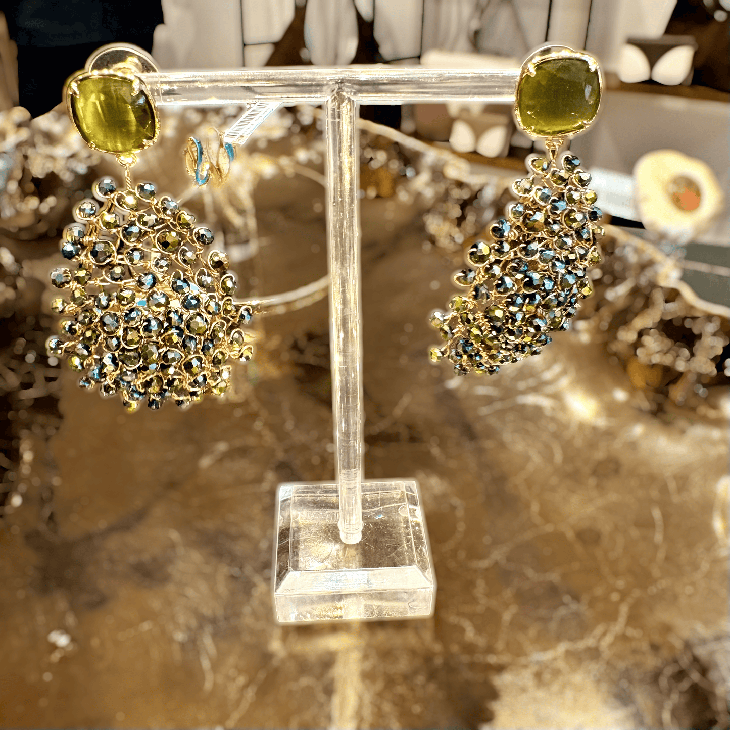 A pair of gold-tone statement earrings displayed on a clear acrylic stand. Each earring features a large oval yellow-green stone at the top, with a dense cluster of small iridescent blue and gold rhinestones below, set against a warm, blurred jewelry display background.