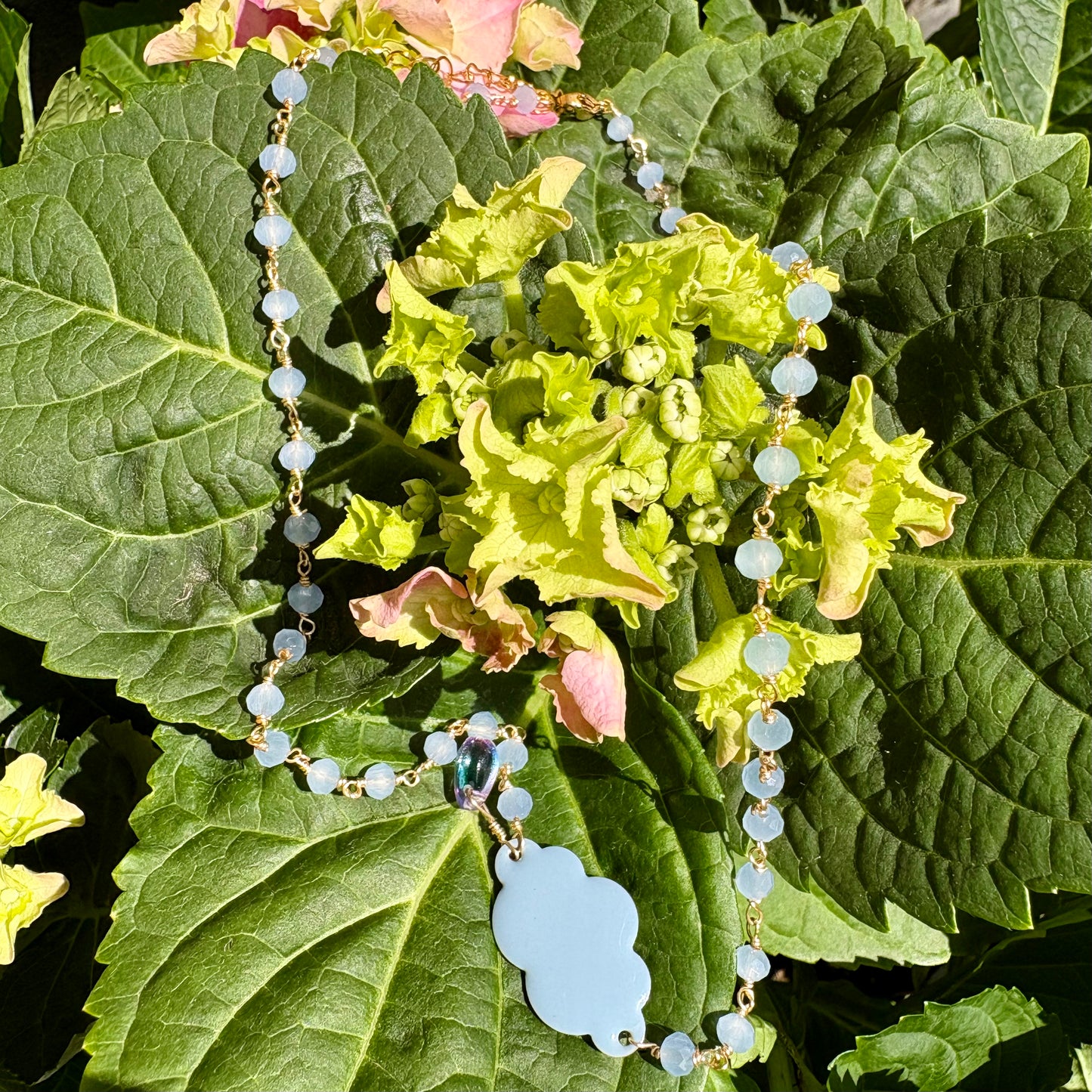 A beaded necklace with light blue faceted beads, gold-tone chain links, and a light blue cloud-shaped pendant, displayed on green hydrangea leaves outdoors.
