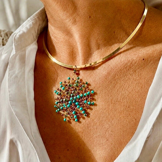 Gold-plated stainless steel choker necklace with a handwoven 18kt gold-plated copper wire Mandàla pendant featuring turquoise and bronze crystal beads, worn over a white linen shirt.