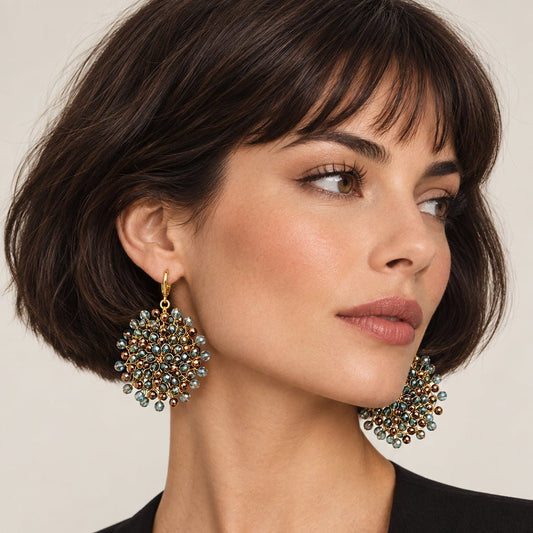 A pair of gold-tone dangle earrings adorned with clusters of small round blue-green stones and darker brown stones worn by a model with a french bob and a black jacket.