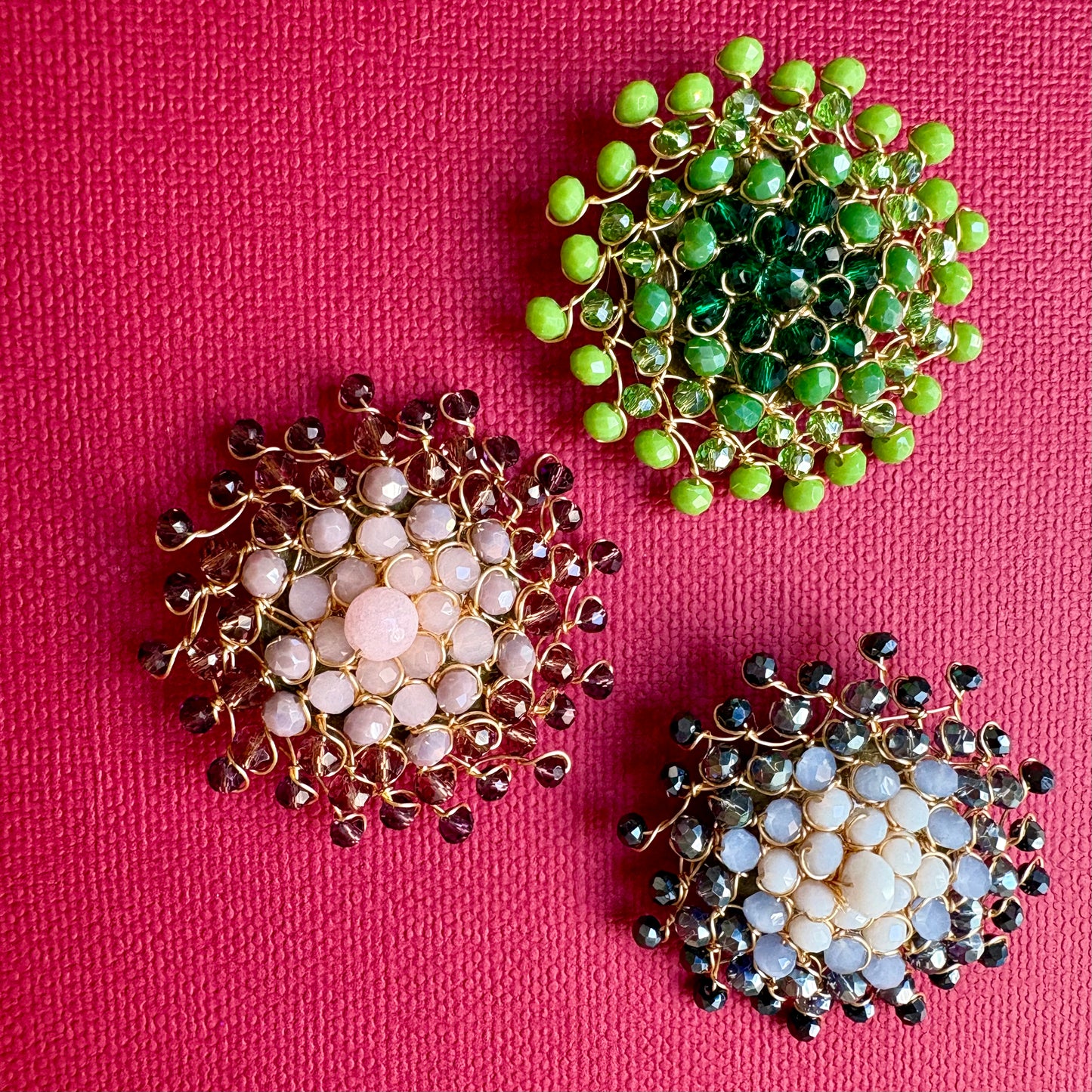 Three handmade gold wire floral brooches with faceted beads, arranged on a textured hot pink background. One brooch features light pink beads, one features dark green beads, and the third features light blue and white beads.