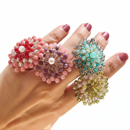 Four handwoven Mandàla rings in red, pink, teal and olive green with champagne crystals, displayed on a hand against a sunny outdoor background.
