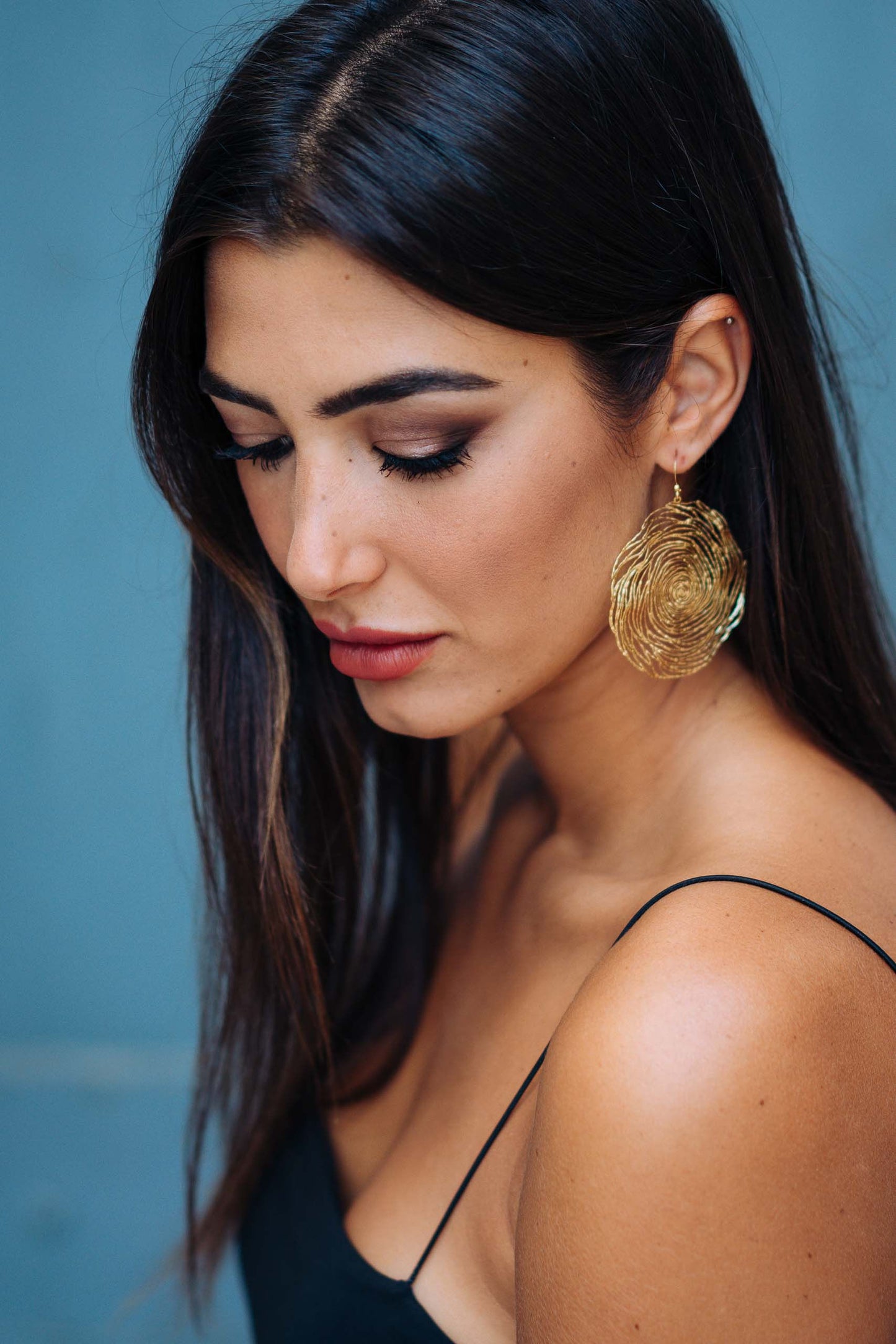 A close-up portrait of a woman with long dark hair wearing a large, round brass wire spiral filigree drop earrings with gold hook fastening