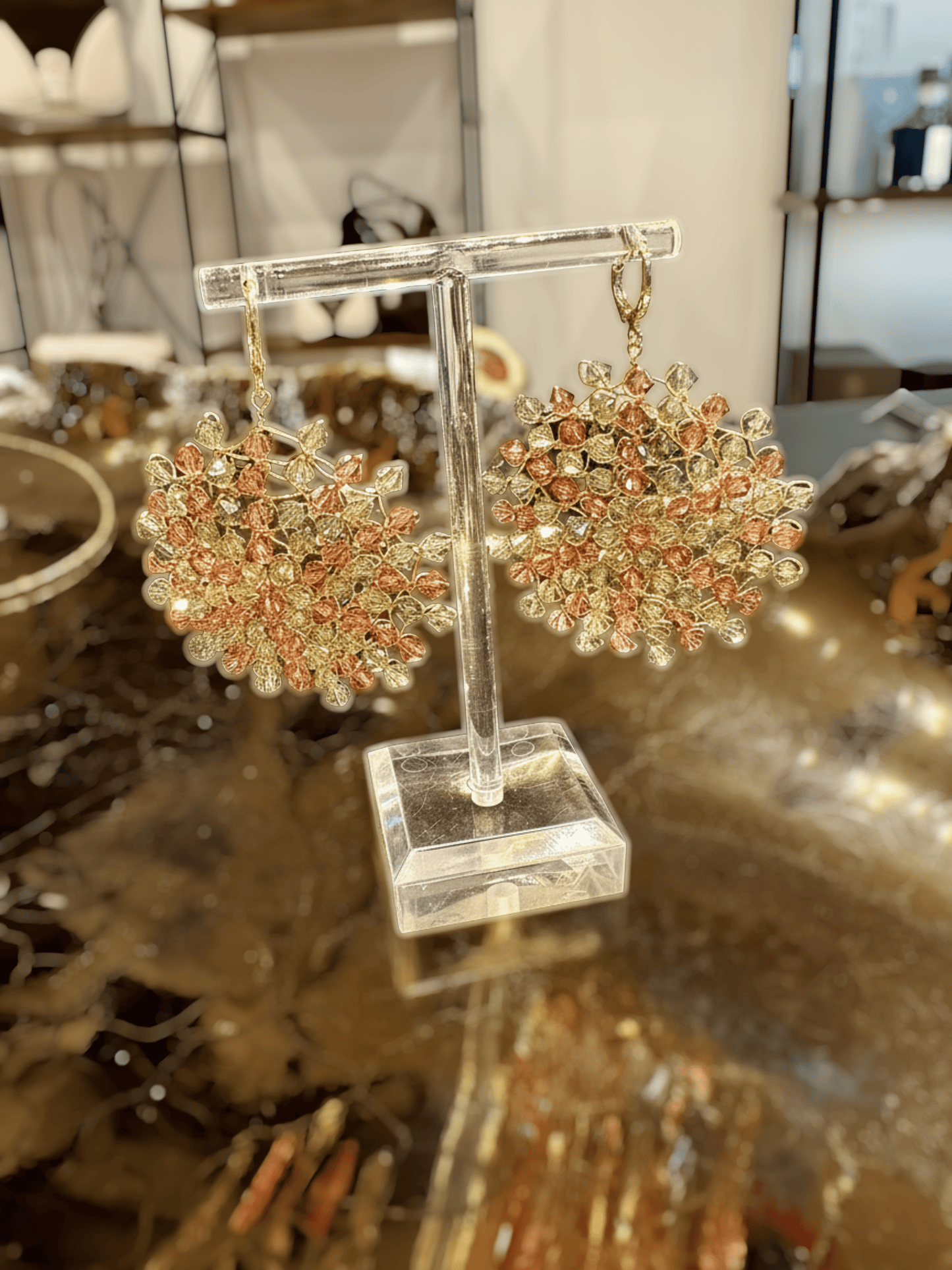 A pair of handmade beaded dangle earrings displayed on a clear acrylic stand. The earrings feature intricate clusters of peach-pink and gold-toned beads with small clear rhinestone accents, hanging from gold-toned hooks.