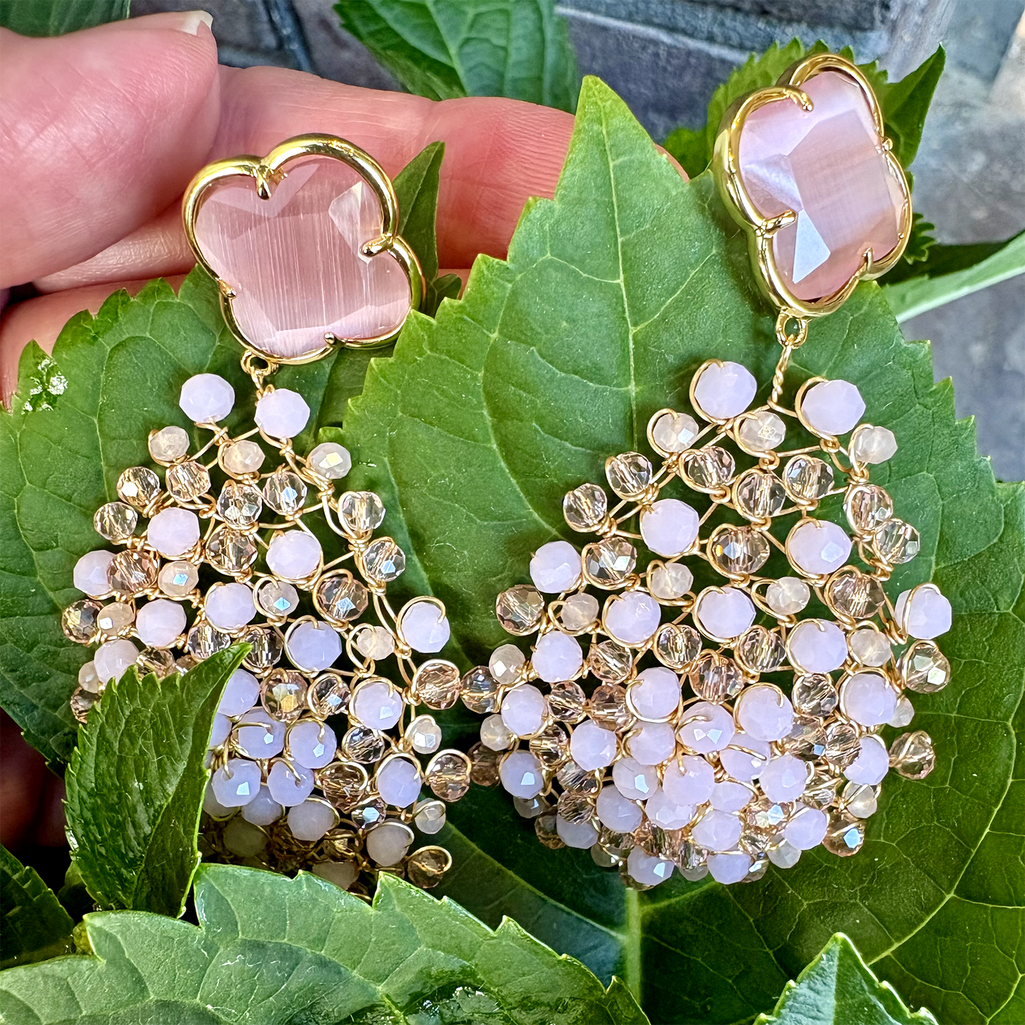 A pair of gold-tone statement dangle earrings with pale pink clover-shaped studs and cascading clusters of clear and white stones, held against large green leaves.