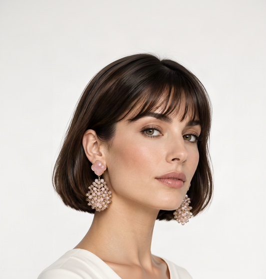 A pair of gold-tone statement dangle earrings with pale pink clover-shaped studs and cascading clusters of clear and white stones on a model with a french bob.