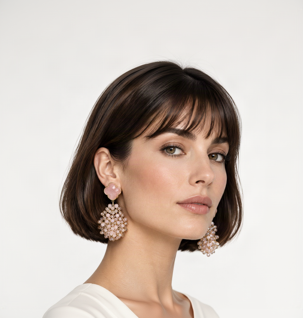 Une paire de boucles d'oreilles pendantes statement dorées avec puces trèfle rose pâle et grappes de pierres transparentes et blanches en cascade, portées par une mannequin au carré court.