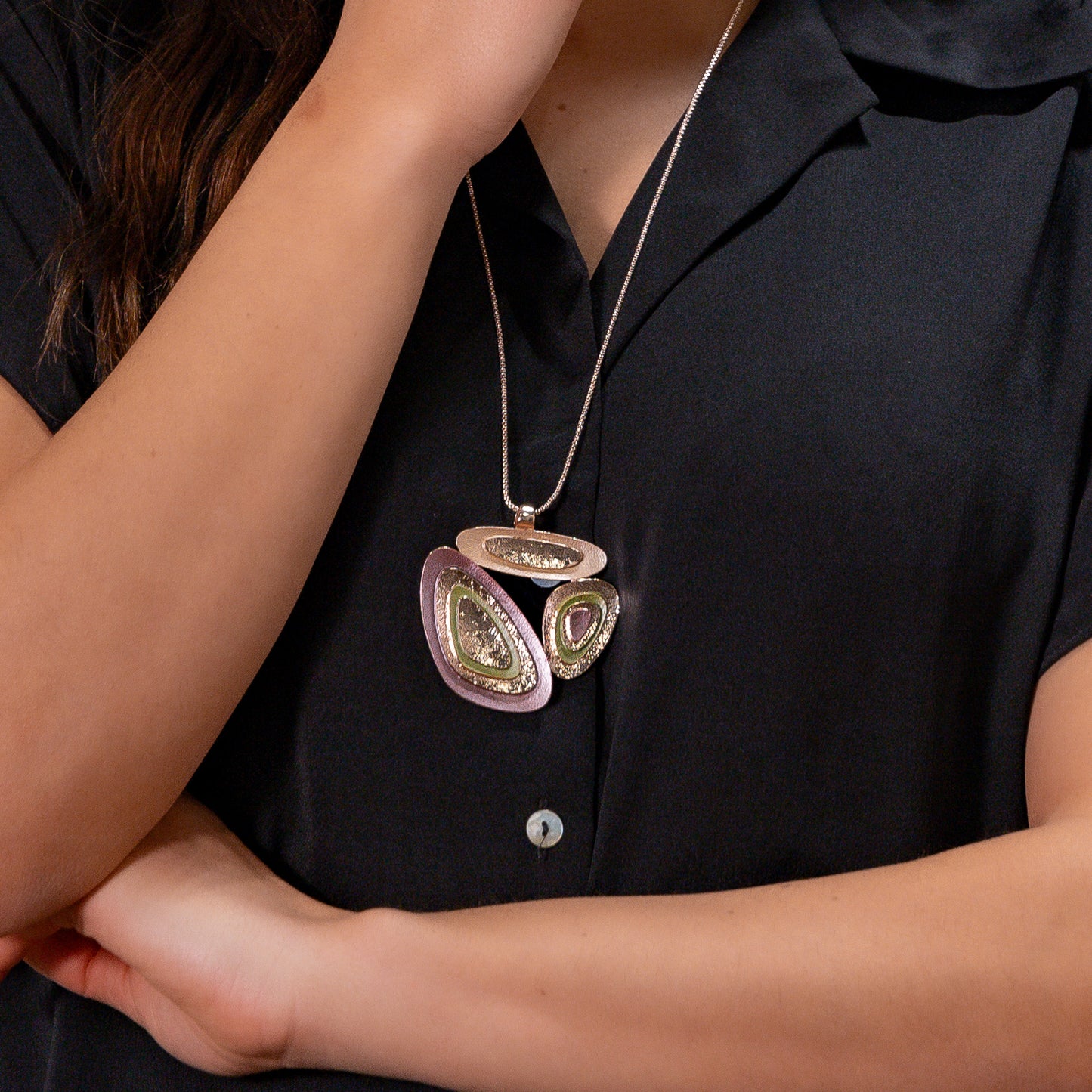 A woman wearing a long rose gold chain necklace with sculptural layered pendant in mauve, sage green and metallic tones on white background