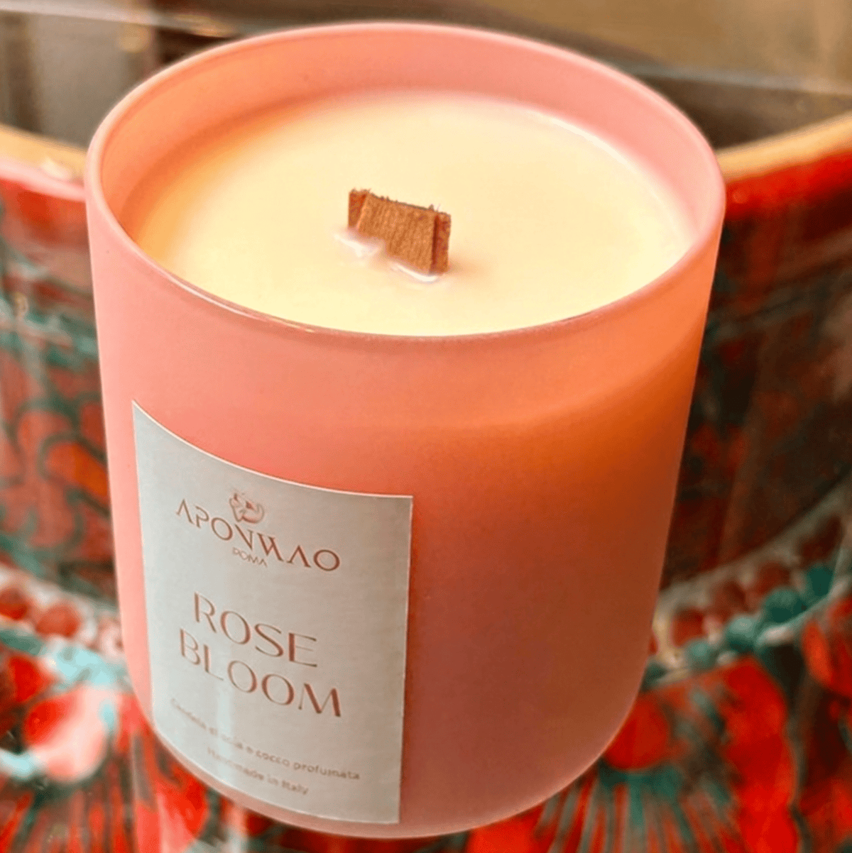 A pink glass jar candle with a white label reading 'APONWAO ROSE BLOOM' and a small wooden wick, set against a colourful patterned background.