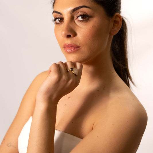Model wearing the Two-Stone Brass Ring with rose quartz and ruby stones, hand raised to chin against a light grey background