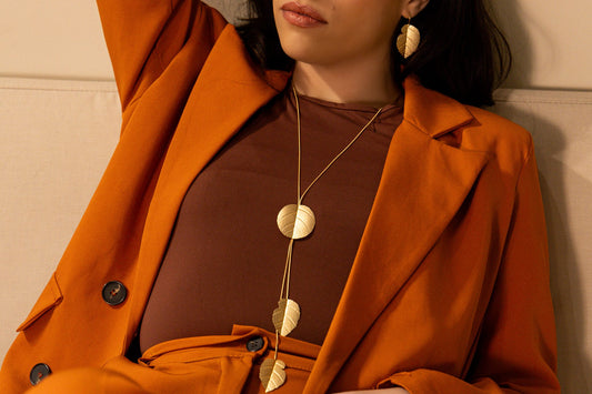Model wearing the Matt Gold Brass Leaf Necklace in matt gold with matching leaf earrings, styled over a brown top and orange blazer