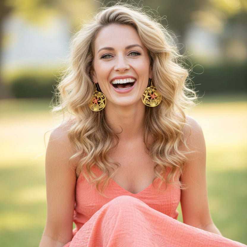 A blonde model wearing a pair of gold pendant earrings with a red star fish and shells. She's laughing. She's wearing a coral coloured sun dress.
