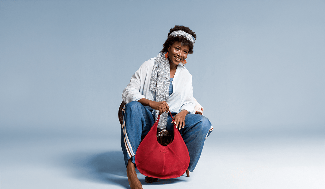 A woman with an afro hairstyle and head scarf sits on a chair, smiling, wearing a light blue linen shirt and relaxed blue jeans while holding a large luxury red suede tote bag.