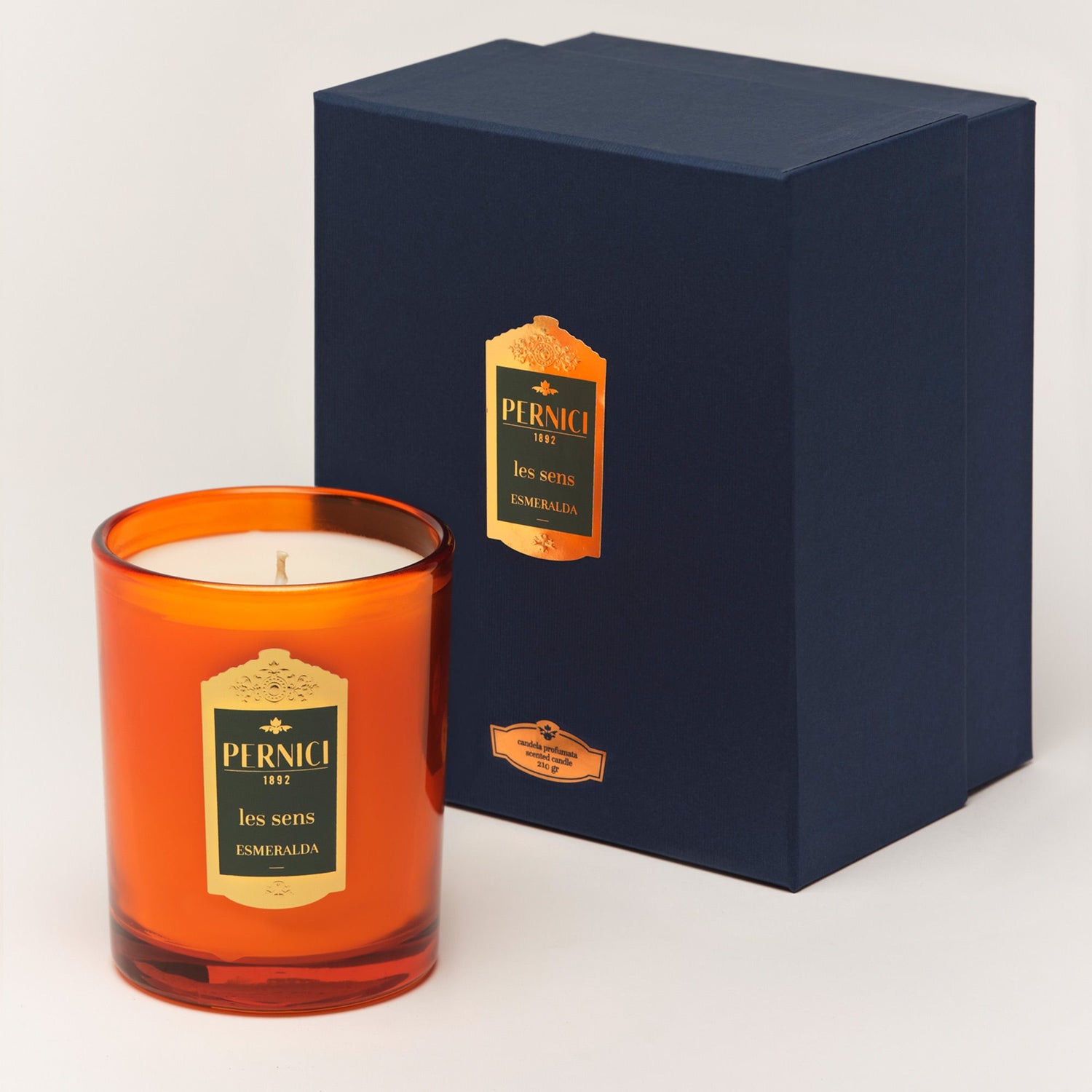 Cereria Pernici Les Sens Esmeralda 210g scented candle in a deep amber glass jar with gold Pernici 1892 label, shown alongside its navy blue gift box with gold foil label, lifestyle shot on warm grey background
