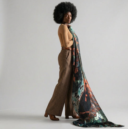 Woman in a camel jumper, checked pleated mini skirt and brown knee-high boots, with the Lys Indigo cashmere scarf draped over her shoulder and trailing to the floor in a wood-panelled hallway with stained-glass windows.