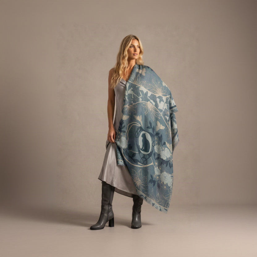 A blonde woman in a grey dress and grey knee-high boots, holding the Leopard Azure cashmere scarf open and draped over one shoulder like a cape, showing the leopard cameo, blue florals and fringed ends against a soft grey backdrop.