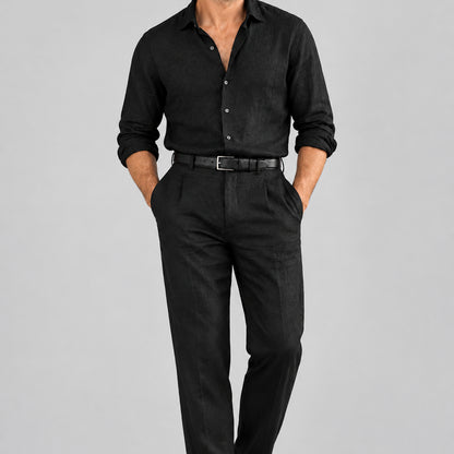 A man wearing a black linen button up shirt and trousers with a black grainy leather belt against a grey background.