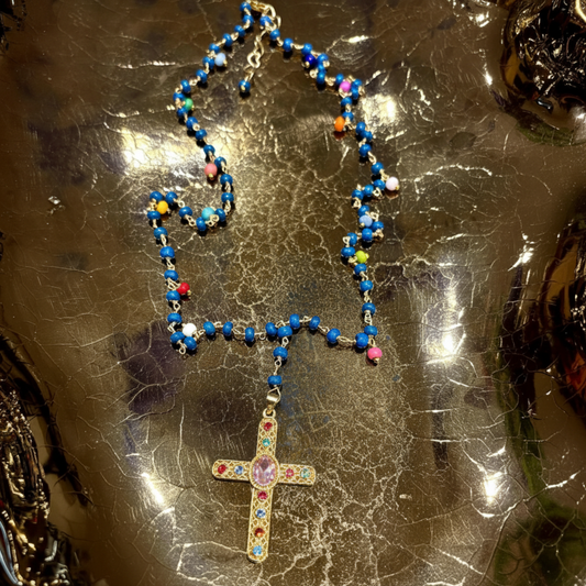 A beaded necklace with a gold-tone cross pendant, featuring predominantly blue beads with small multicolored accents, displayed on a reflective metallic surface.