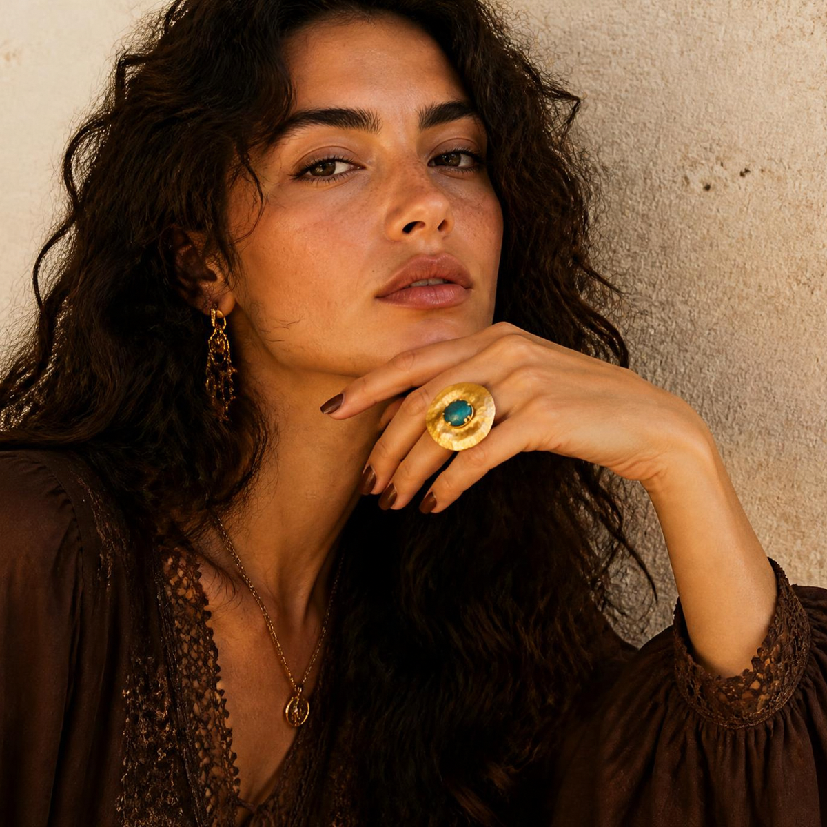 A woman with long, wavy dark hair wearing a brown bohemian-style top and gold jewelry, including a large gold ring with a blue and green stone, posing against a textured beige plaster wall.