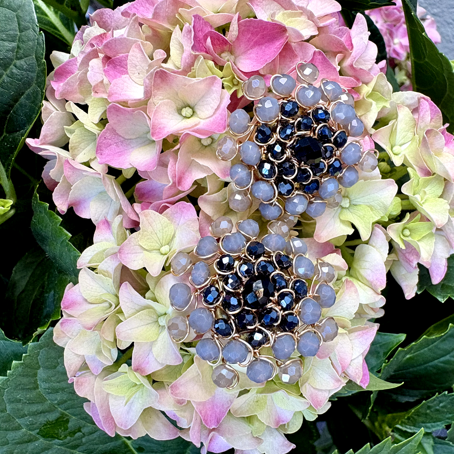 A pair of button covers featuring rows of faceted light blue-gray beads and dark blue center stones, displayed on a pink and green hydrangea flower.
