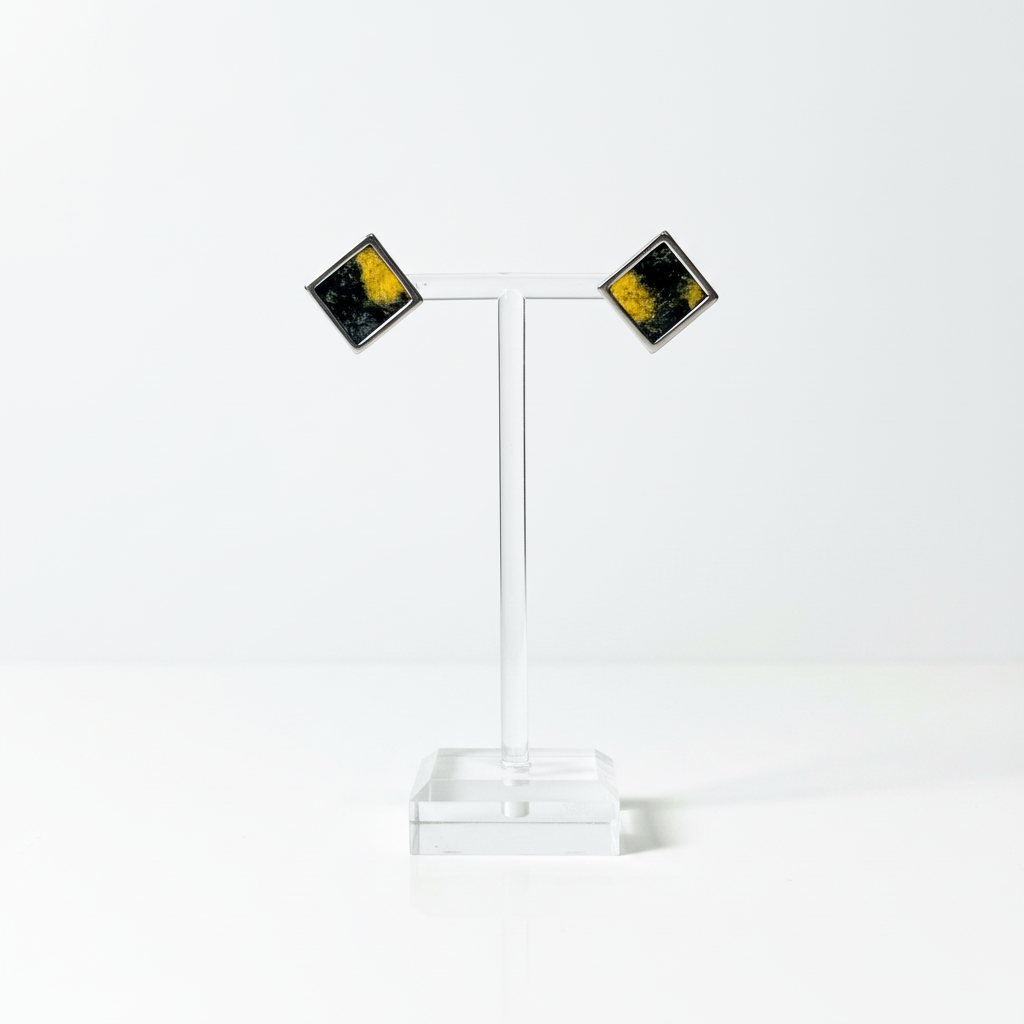 A pair of square stud earrings with a silver-tone border and a black and yellow marbled pattern in Nuno Felt, displayed on a clear acrylic stand on a white background.