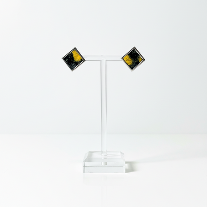 A pair of square stud earrings with a silver-tone border and a black and yellow marbled pattern in Nuno Felt, displayed on a clear acrylic stand on a white background.