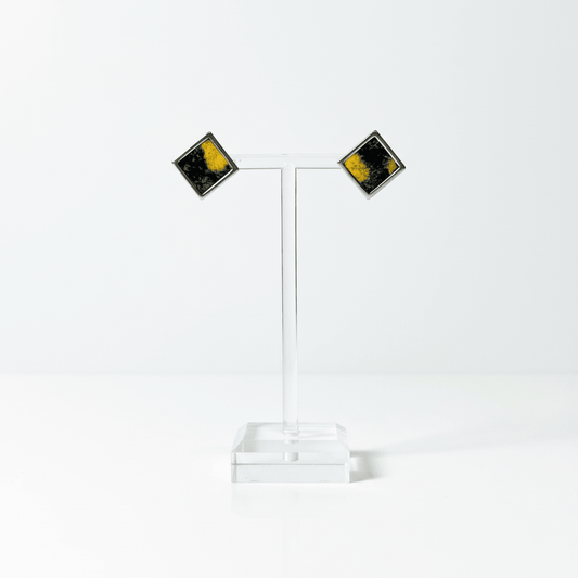 A pair of square stud earrings with a silver-tone border and a black and yellow marbled pattern in Nuno Felt, displayed on a clear acrylic stand on a white background.