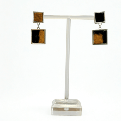 A pair of square drop earrings displayed on a clear acrylic T-stand against a white background. Each earring features a top silver-tone square with a black and ocre marbled pattern, connected by a small silver chain to a larger bottom square of the same design.