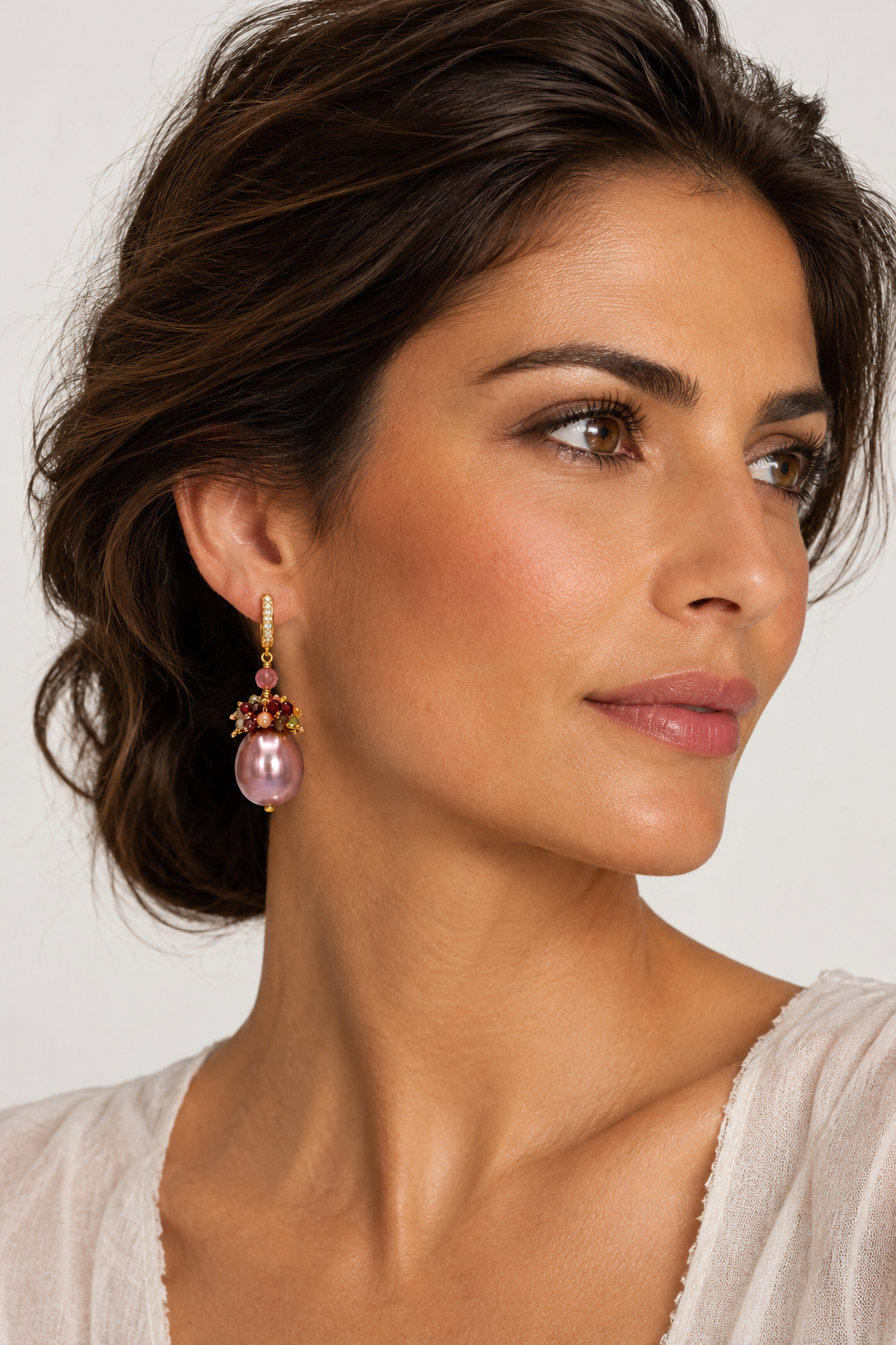 A close-up of a woman wearing a dangling earring with a pink Maiorca pearl, gold-toned metal, and coloured tourmaline accents against a light neutral background.