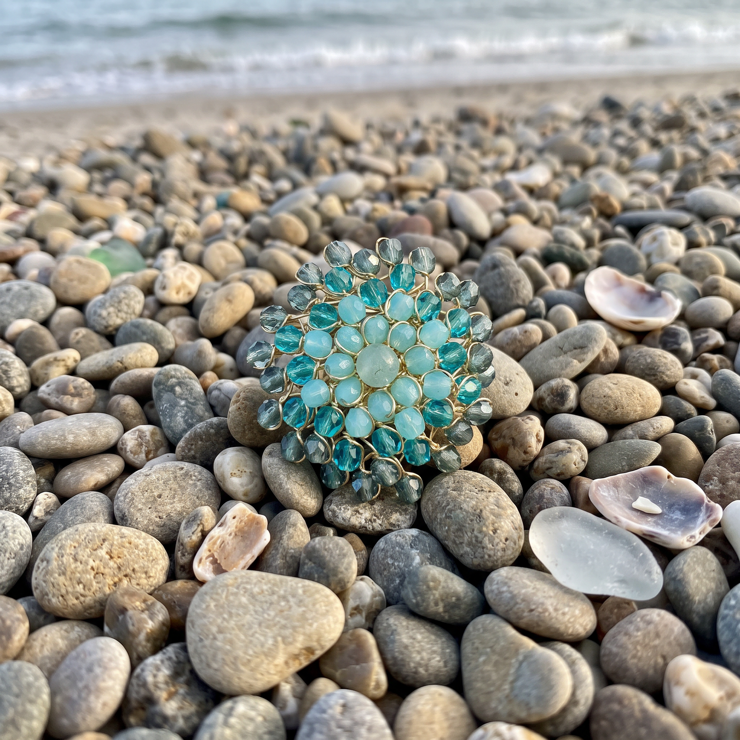 A round gold-tone brooch with turquoise-blue faceted gemstones, resting on a beach of smooth gray and beige pebbles with the shoreline and gentle waves blurred in the background.