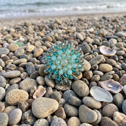 A round gold-tone brooch with turquoise-blue faceted gemstones, resting on a beach of smooth gray and beige pebbles with the shoreline and gentle waves blurred in the background.