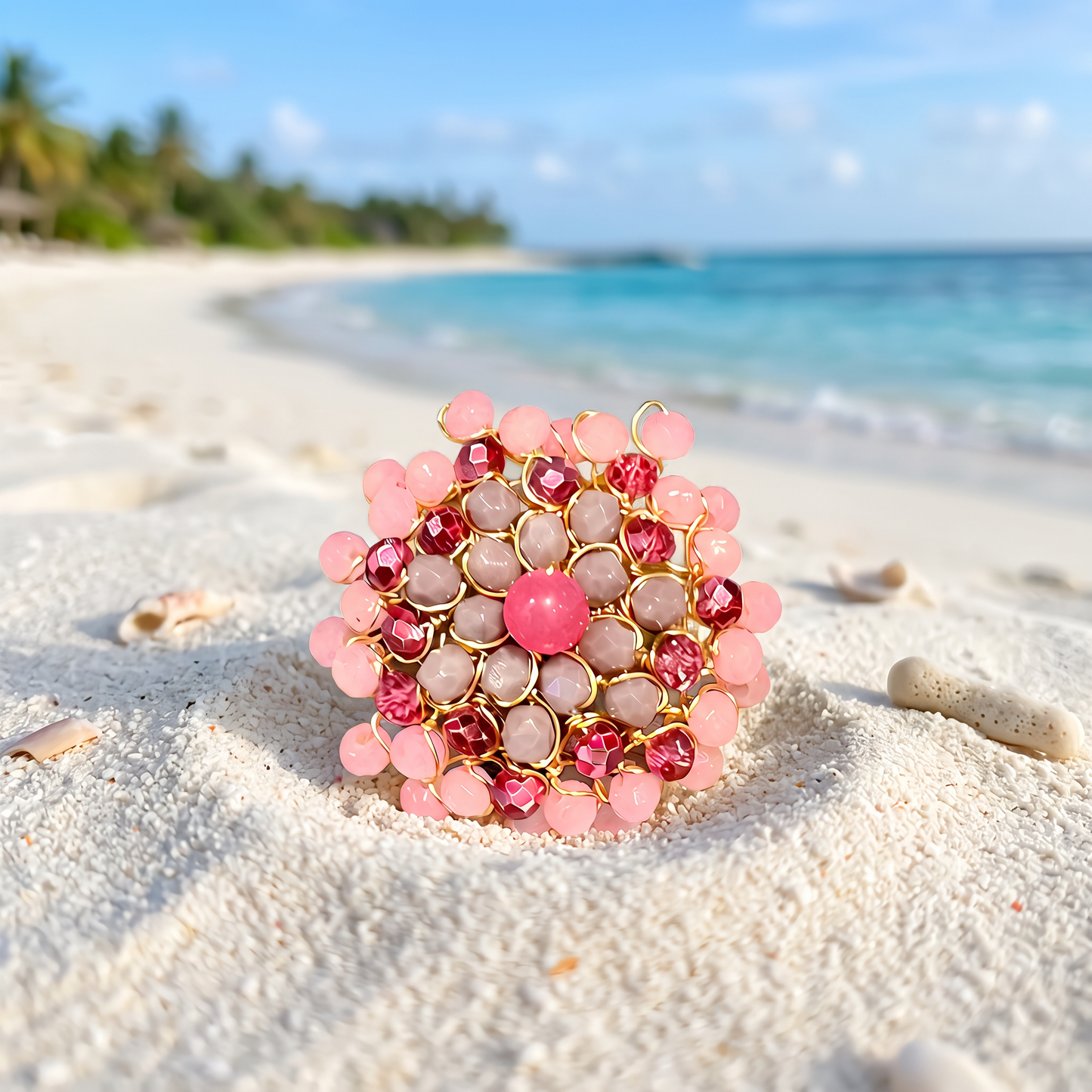 A pink and gold beaded brooch with faceted crystal accents, displayed on a white sand beach with a tropical ocean and palm trees in the background.