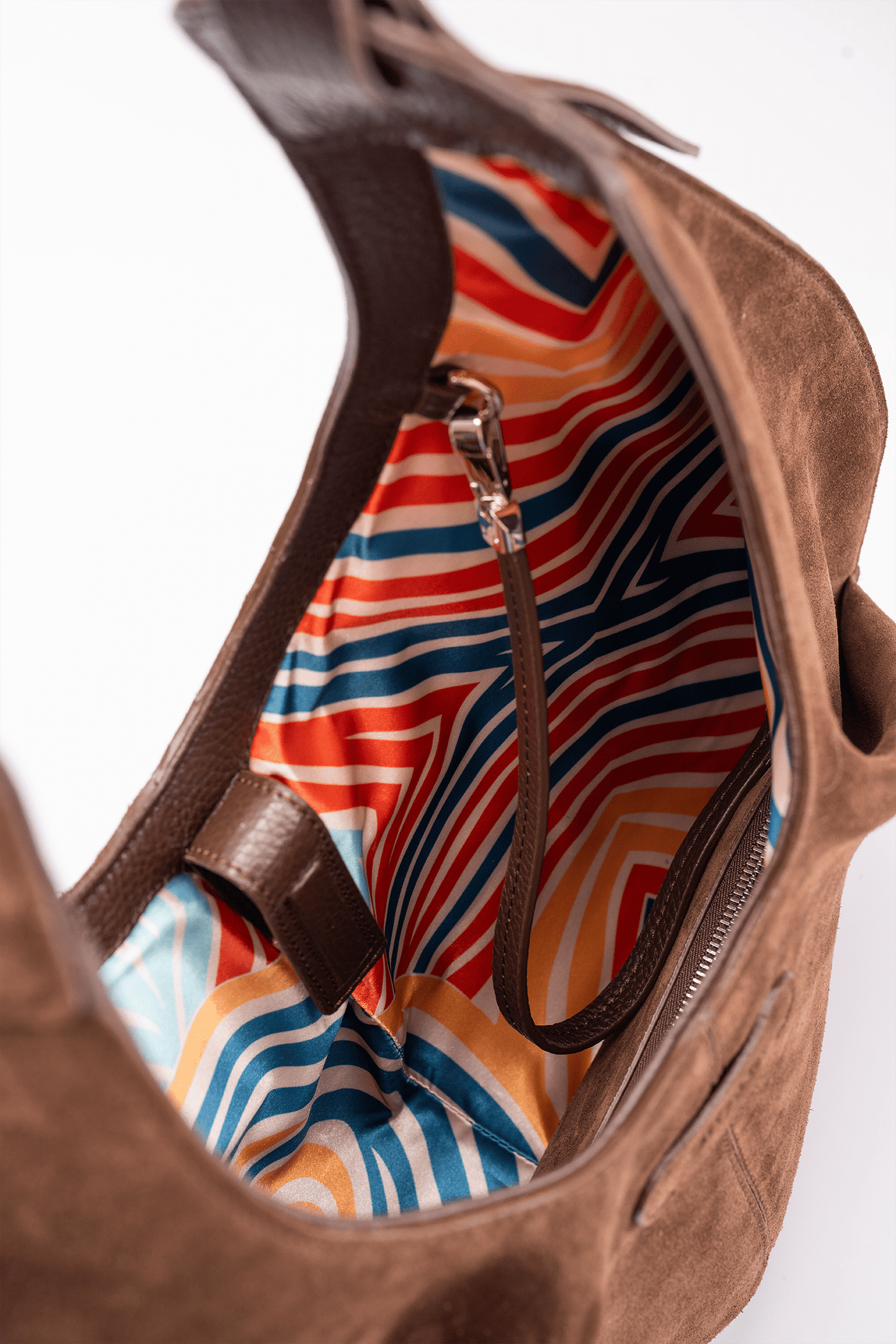 Top view of the Gitana suede shoulder bag in warm brown leather, showcasing the spacious interior with vibrant geometric striped lining, zip closure, and detachable inner leather pouch strap detail.
