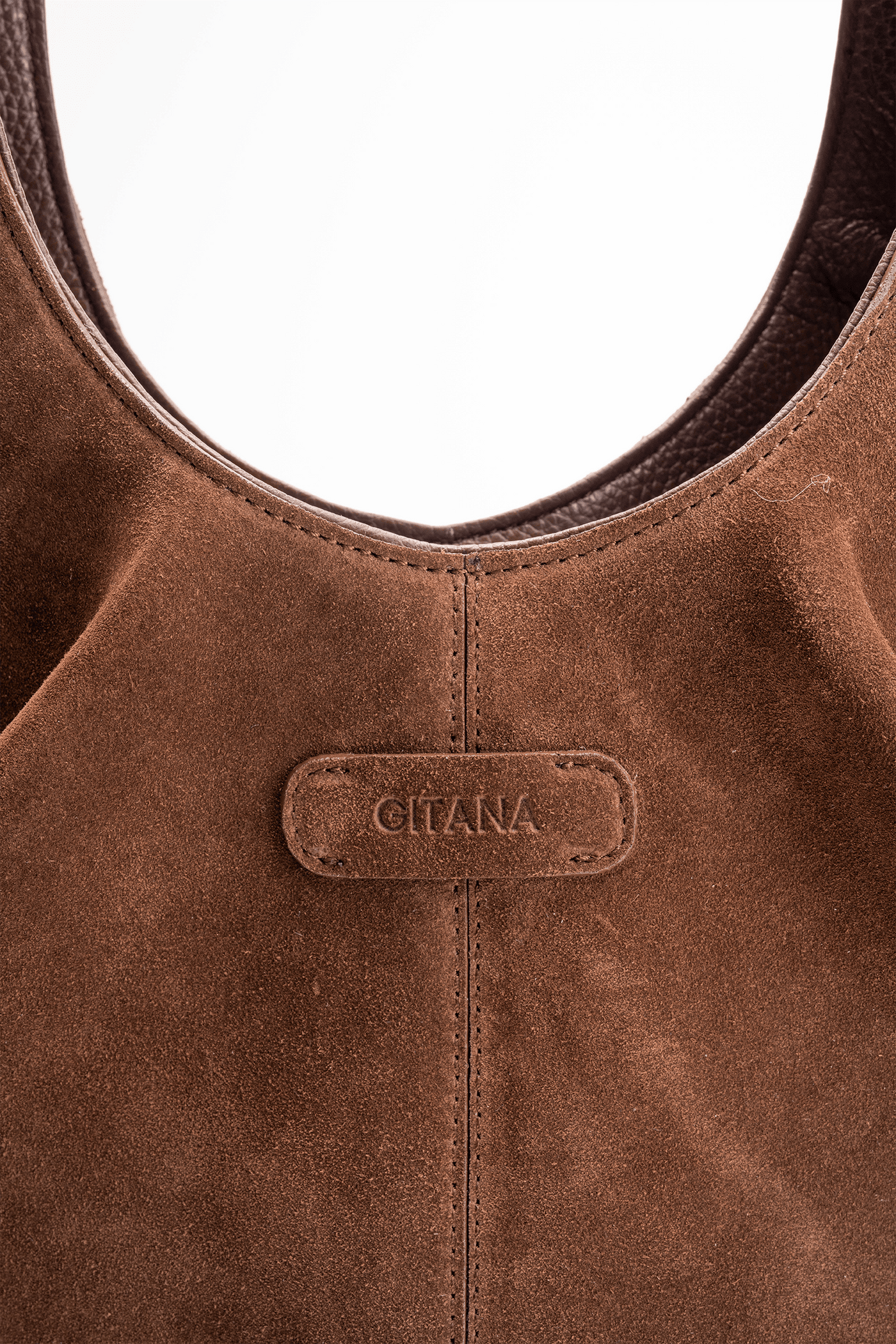 Close-up detail of the Gitana suede shoulder bag in warm  brown leather, featuring the embossed Gitana logo, curved top handle, contrast stitching, and soft textured suede finish.