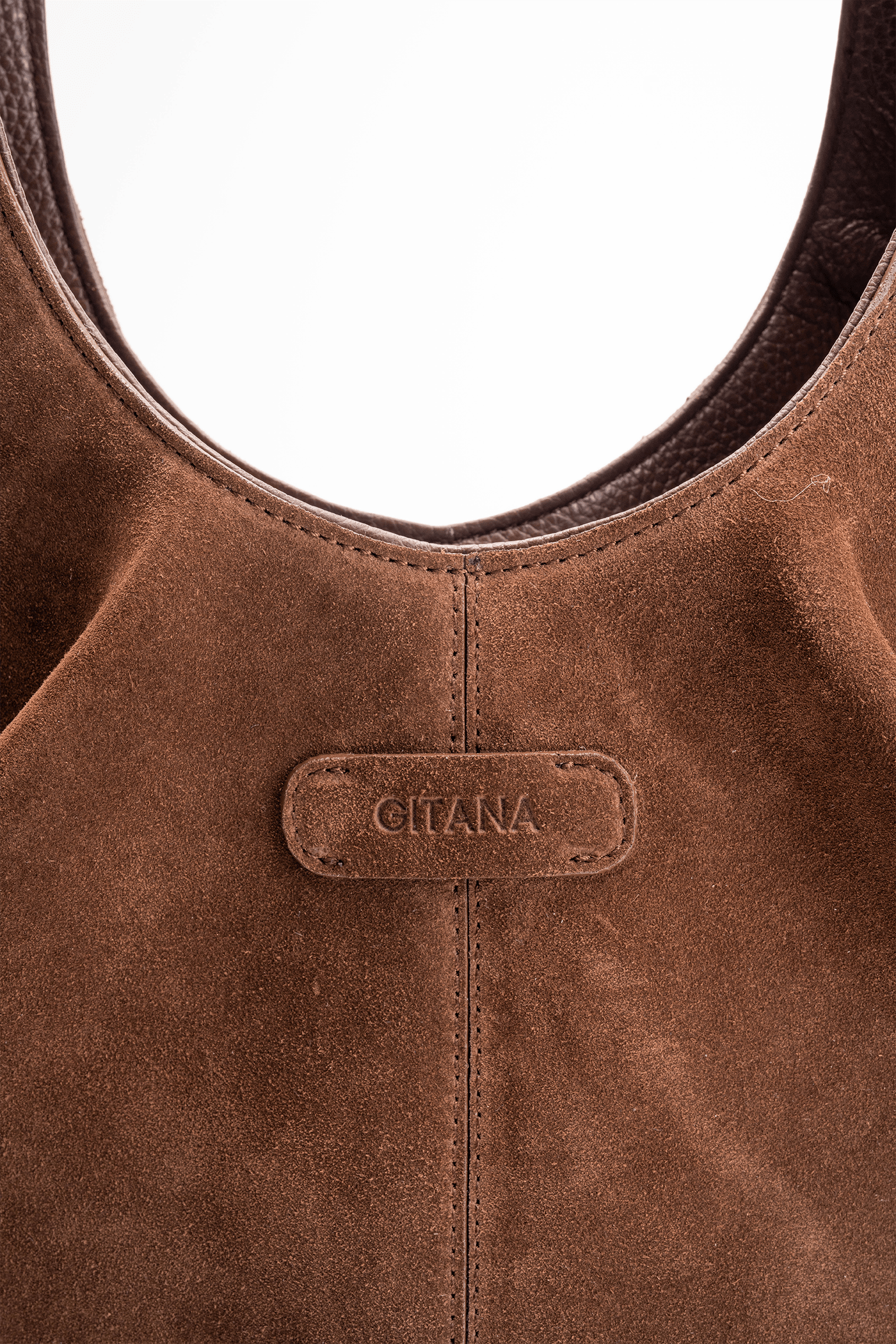 Close-up detail of the Gitana suede shoulder bag in warm  brown leather, featuring the embossed Gitana logo, curved top handle, contrast stitching, and soft textured suede finish.