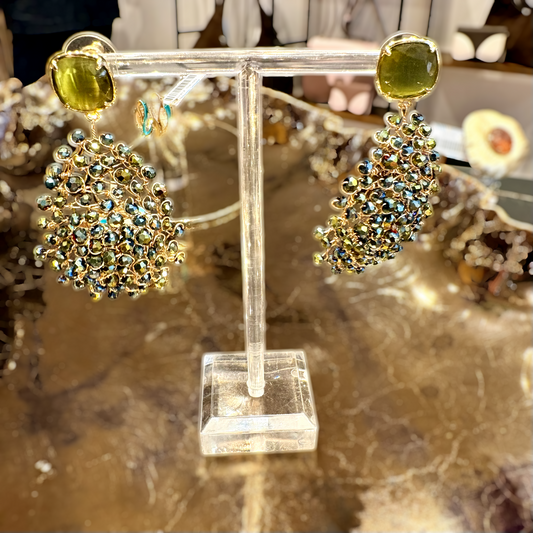 A pair of gold-tone statement earrings displayed on a clear acrylic stand. Each earring features a large oval yellow-green stone at the top, with a dense cluster of small iridescent blue and gold rhinestones below, set against a warm, blurred jewelry display background.