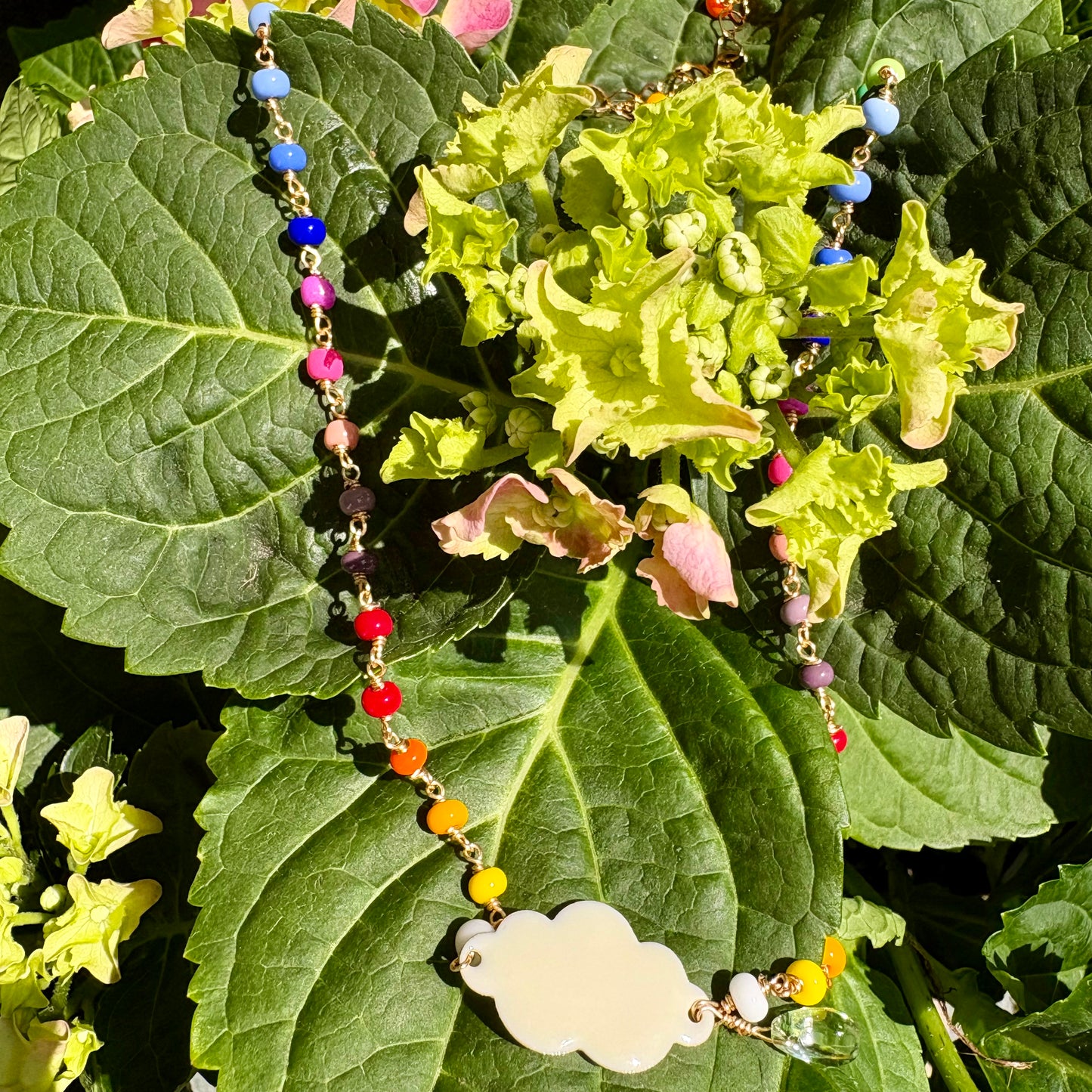 A colorful beaded necklace with a blank white cloud-shaped pendant, draped over green hydrangea leaves in natural outdoor light.