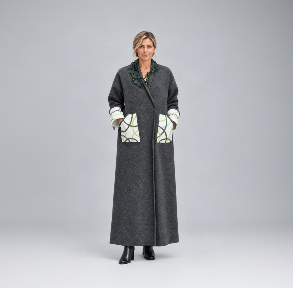 A woman wearing a long charcoal gray wool-blend coat with large patch pockets featuring a white, green, and black abstract pattern, paired with a chunky green necklace.