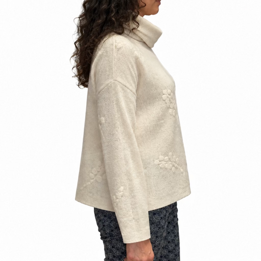 The side view of a woman wearing a cream merino wool cowl neck top with a cream grape design and patterned blue pants.