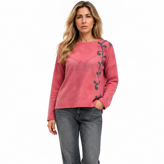 A woman wearing a pink merino wool crew neck with a fleur d'alis grey pattern down the front side.  She's wearing Jeans.