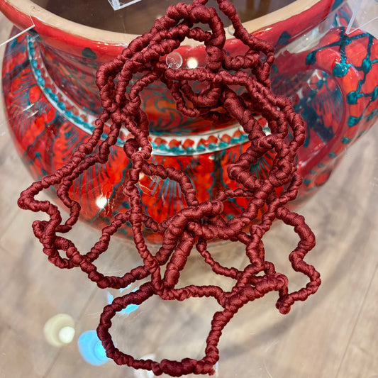 A close-up of a coiled, frizzy, deep red synthetic fiber or yarn draped over a large, glossy red ceramic pot with blue and black floral patterns, set on a light wood floor.