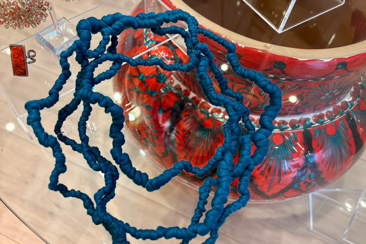 A chunky, textured blue beaded necklace draped over a clear acrylic display stand, with a red and teal patterned bowl visible in the background.